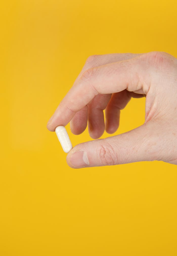 A close-up of a hand holding a white capsule against a vibrant yellow background.